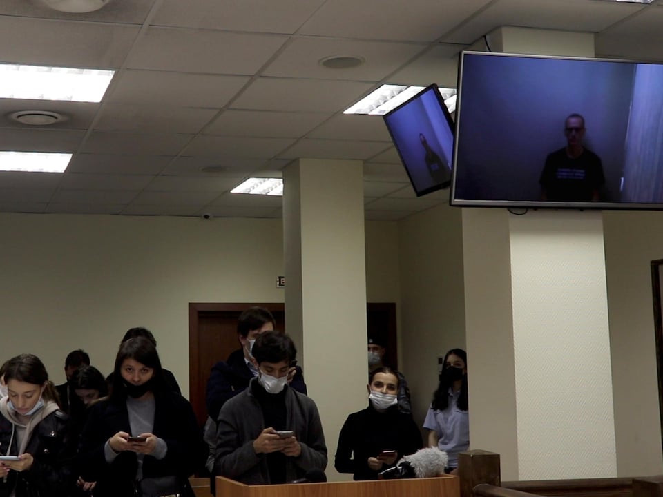 People wearing face masks look at their cell phones in the courtroom.  Screens show Alexei Navalny.