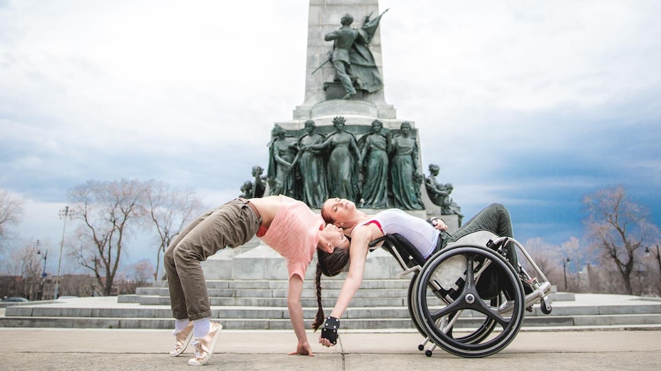 Dancers Joannie Douville and France Geoffroy (to the right, in a wheelchair) lean back to do dances next to a pole with many statues.