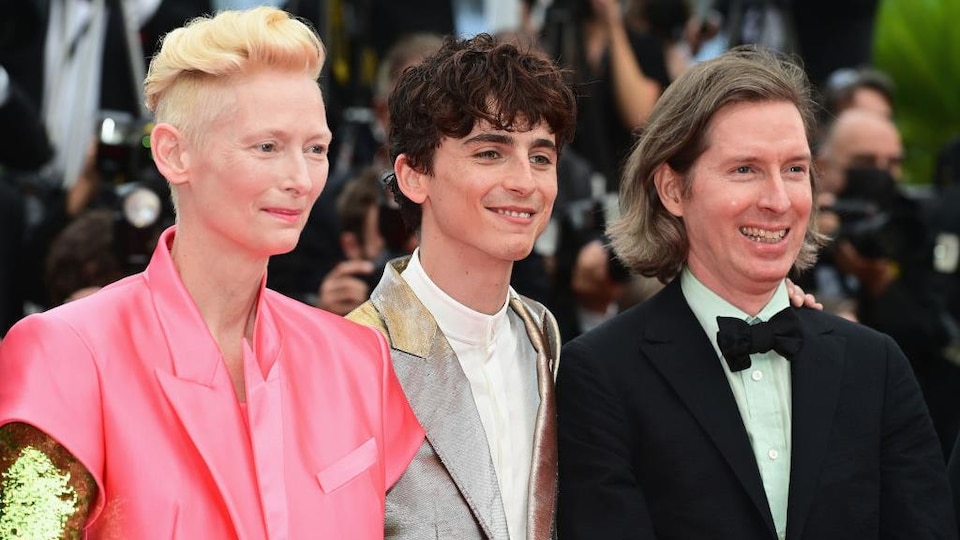 Tilda Swinton wears a pink jacket, Timothée Chalamet wears a gray satin jacket, and Wes Anderson wears a black jacket.