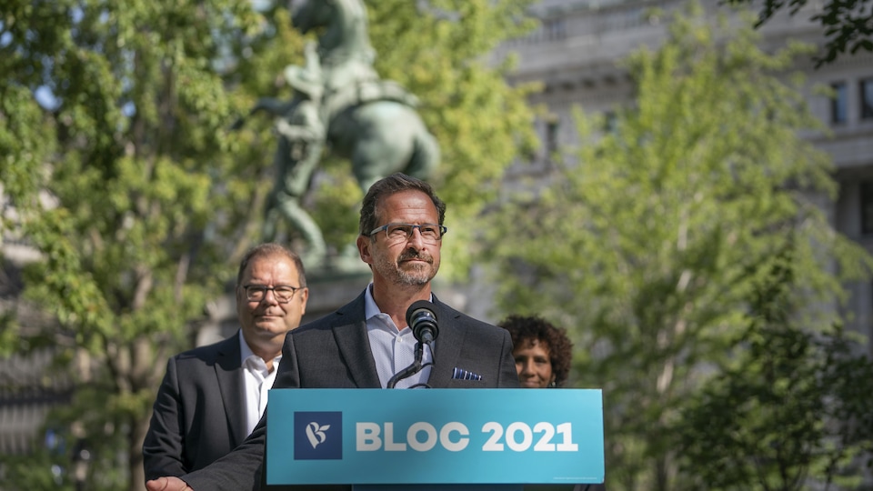 Mr Blanchett speaks to the media, standing face to face.  Behind him stand Mr. Beaulieu and Mrs. Michel.  In the background, the statue, trees, and buildings surrounding the Place du Canada in Montreal.