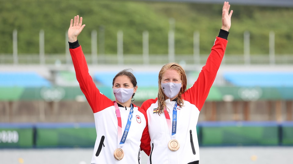 Two masked Canadian women with bronze medals around their necks holding each other at the waist and waving with one hand.