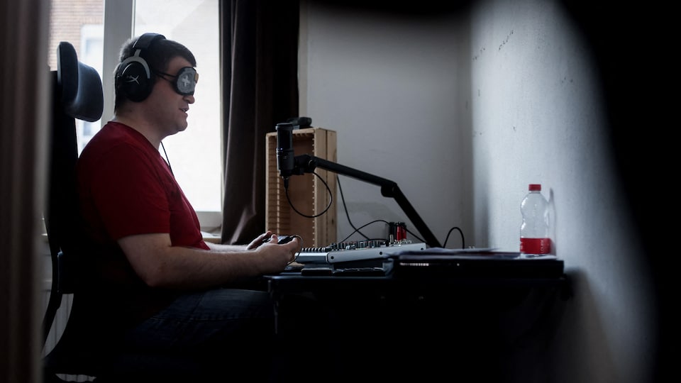 The man is blindfolded and with headphones, holding the console and sitting at his desk in front of a white wall without a screen.