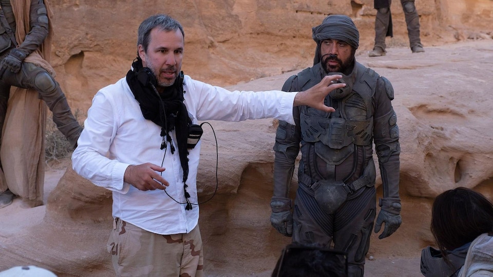 A man in a white shirt gives directions to others in a desert scene.