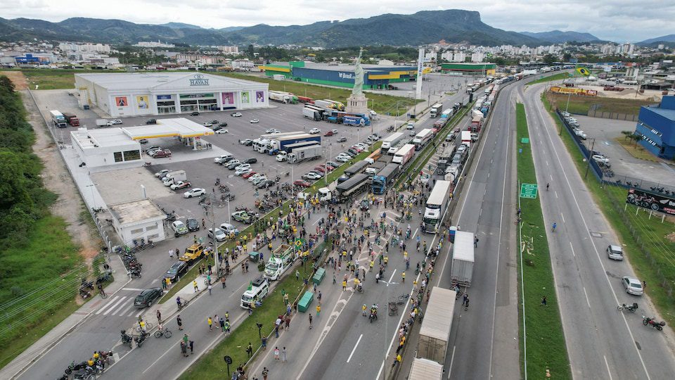 People are walking in the middle of a highway blocked by heavy trucks.